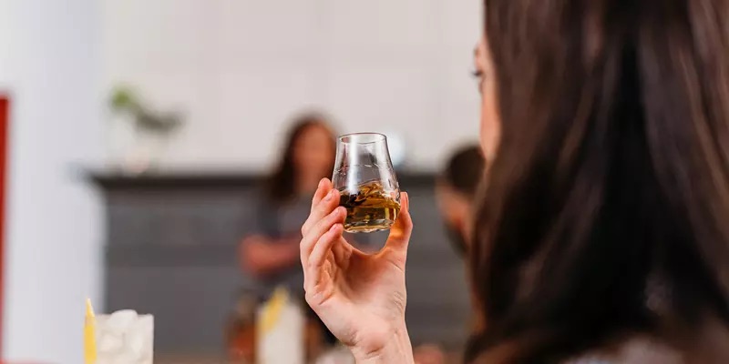 A person with long brown hair holds a glass of whiskey, with blurred figures and a table visible in the background.