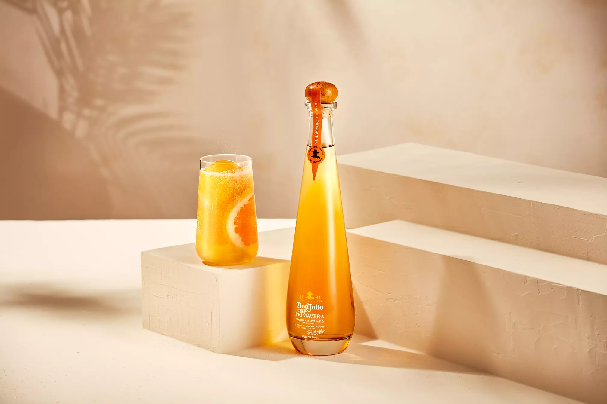A tall bottle of Don Julio Primavera tequila stands next to a glass of orange cocktail with an orange slice, set on white geometric platforms.