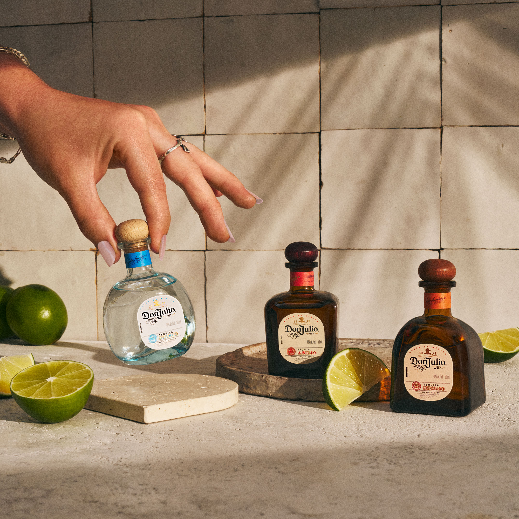 Hand reaching for a small bottle of Don Julio Blanco tequila, with Don Julio Reposado and Añejo bottles, lime halves, and a tiled background.