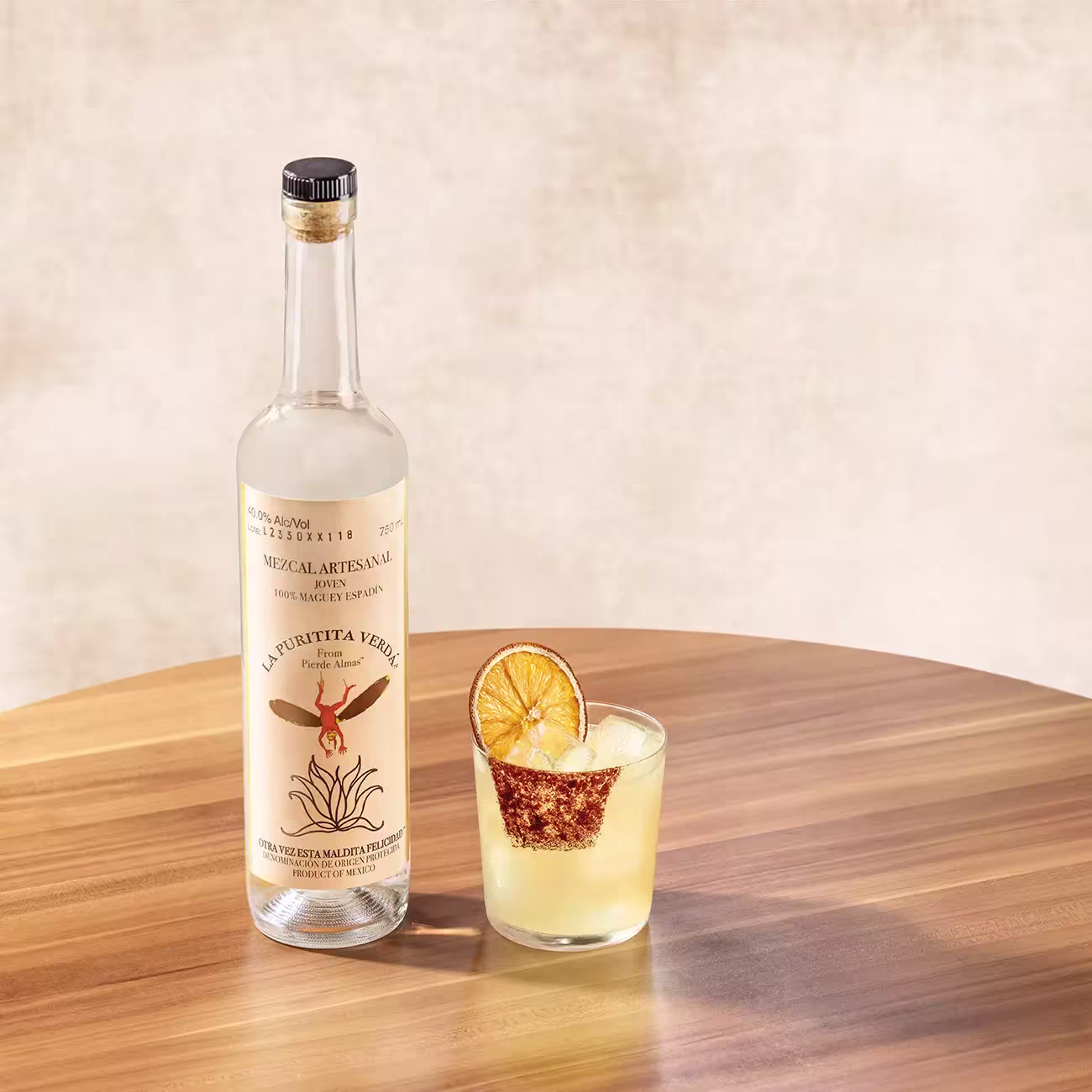 A bottle of mezcal and a cocktail garnished with dried orange sit on a round wooden table against a neutral background.