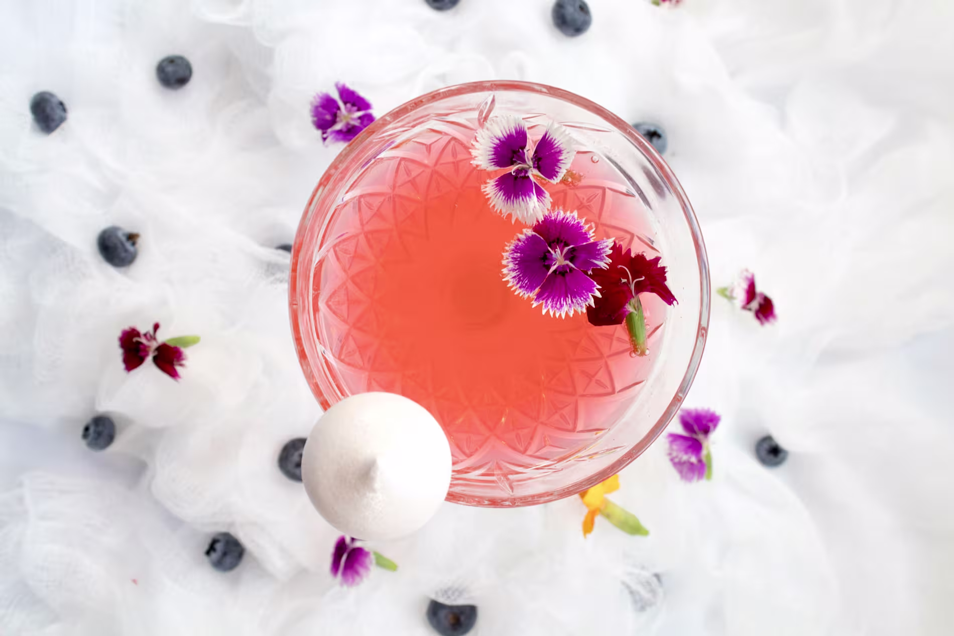 A pink beverage in a patterned glass, garnished with purple and white flowers, sits on a white cloth with scattered blueberries and a white meringue.