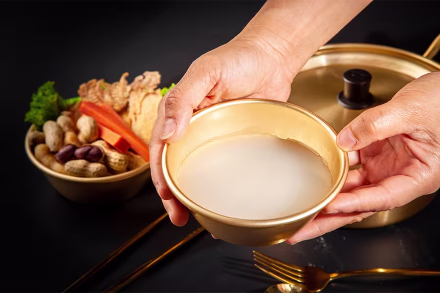 A person holds a brass bowl filled with a milky white liquid; a bowl of assorted nuts and vegetables and a brass pot are in the background.