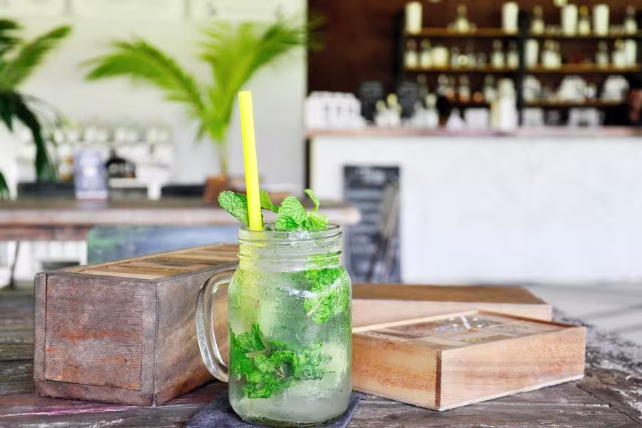 A mason jar filled with a mint beverage, ice, and a yellow straw sits on a wooden table indoors, with leafy plants and shelves in the blurred background.