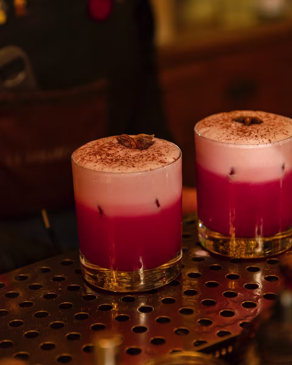 Un primer plano de cócteles de La Confitería con tonos rojizos y una espuma rosada decorada con anís estrellado, sobre una barra metálica.