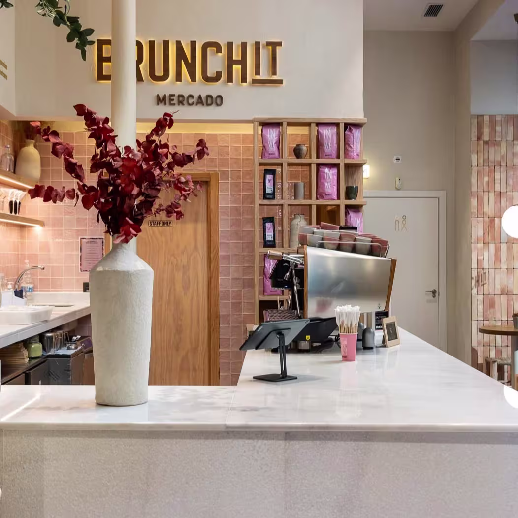 Mostrador de barra con superficie blanca, detalles en tonos tierra y rosas, y decoración floral, bajo el rótulo dorado de 'Brunchit Mercado', en un interior cálido y moderno.