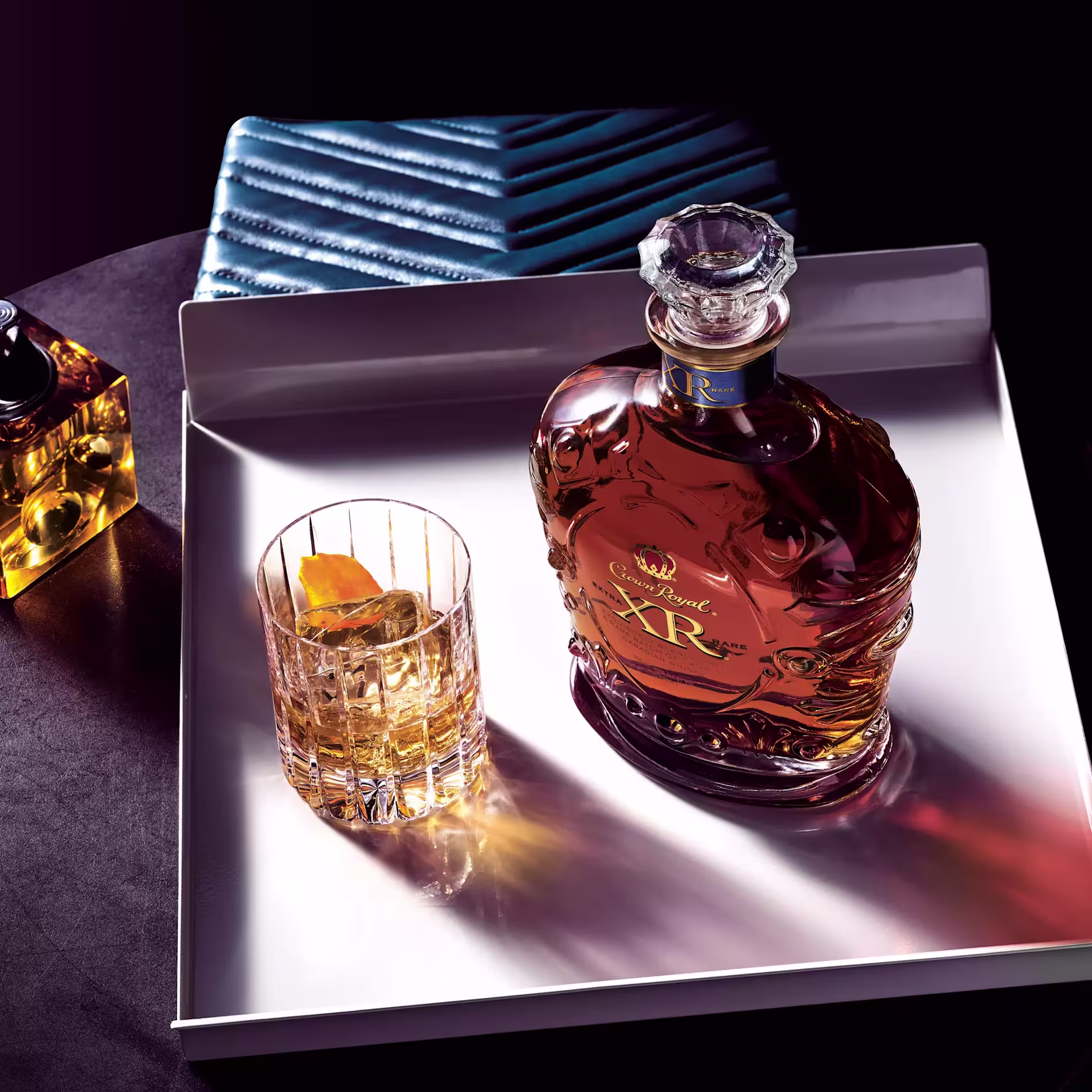 A bottle of Crown Royal XR whisky and a glass with whisky and an orange peel sit on a white tray next to a blue textured case.