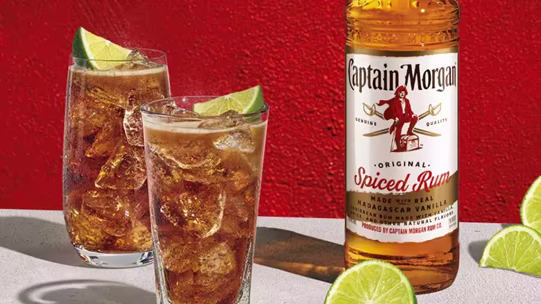 A bottle of Captain Morgan Original Spiced Rum stands next to two glasses filled with a dark beverage, ice, and lime wedges, with extra lime slices on a white surface against a red wall.