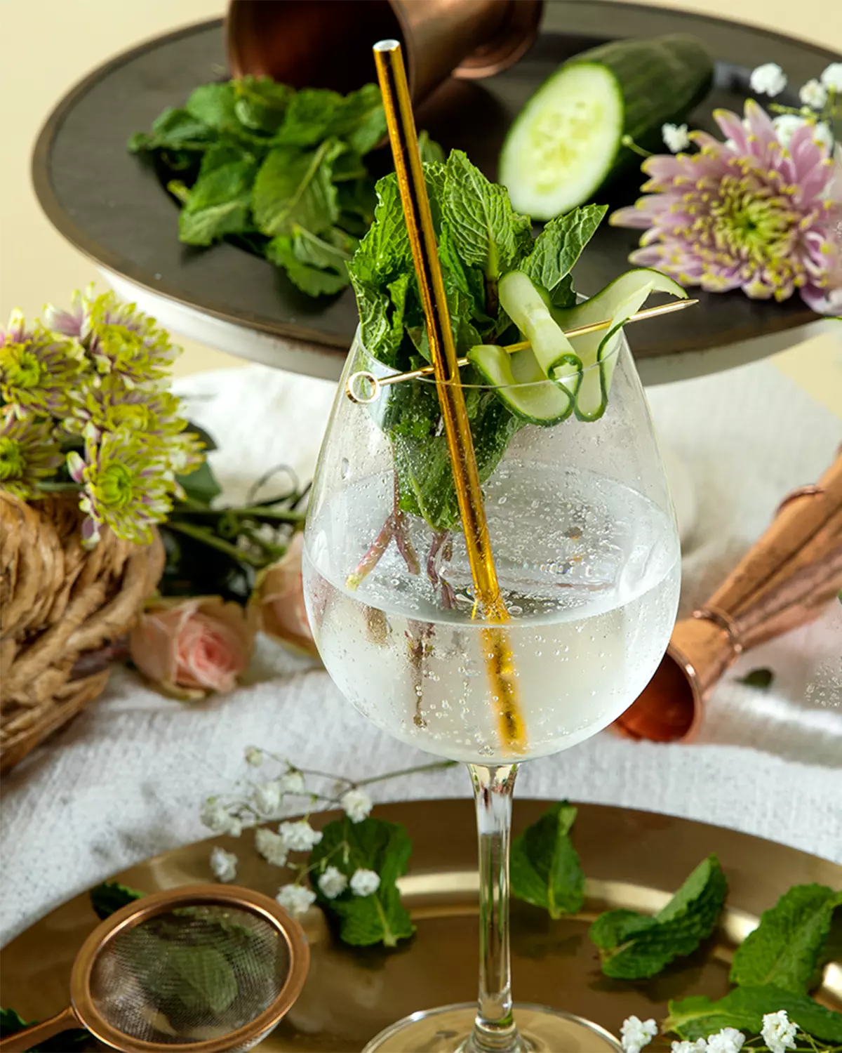 A glass of sparkling water garnished with fresh mint and a cucumber ribbon, with a gold straw, surrounded by flowers, mint leaves, and cocktail tools on decorative trays.