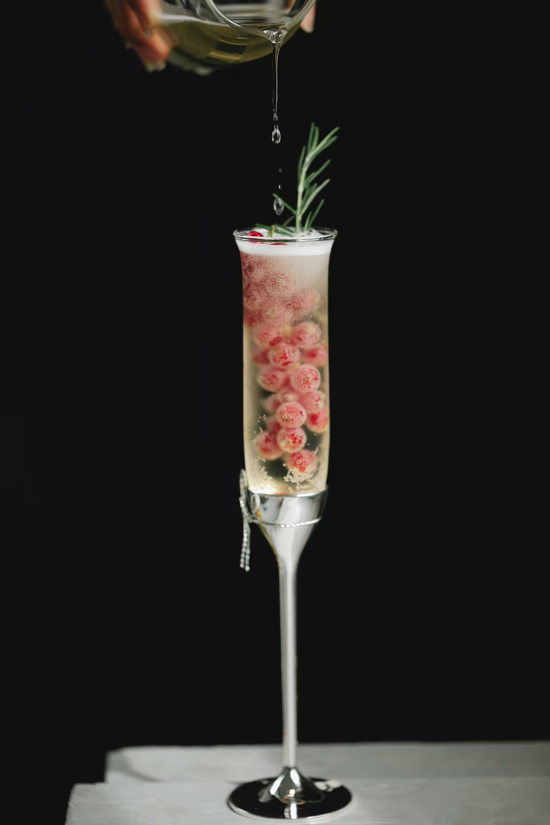 A champagne flute filled with light-colored beverage, floating red berries, and a rosemary sprig, with liquid being poured from above against a black background.