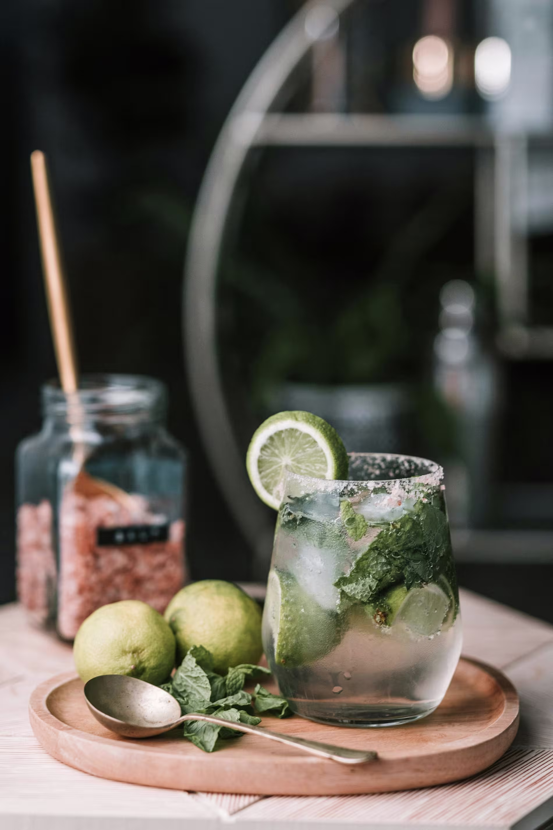 A glass of mojito with lime and mint sits on a wooden tray beside whole limes, a spoon, and a jar of brown sugar.
