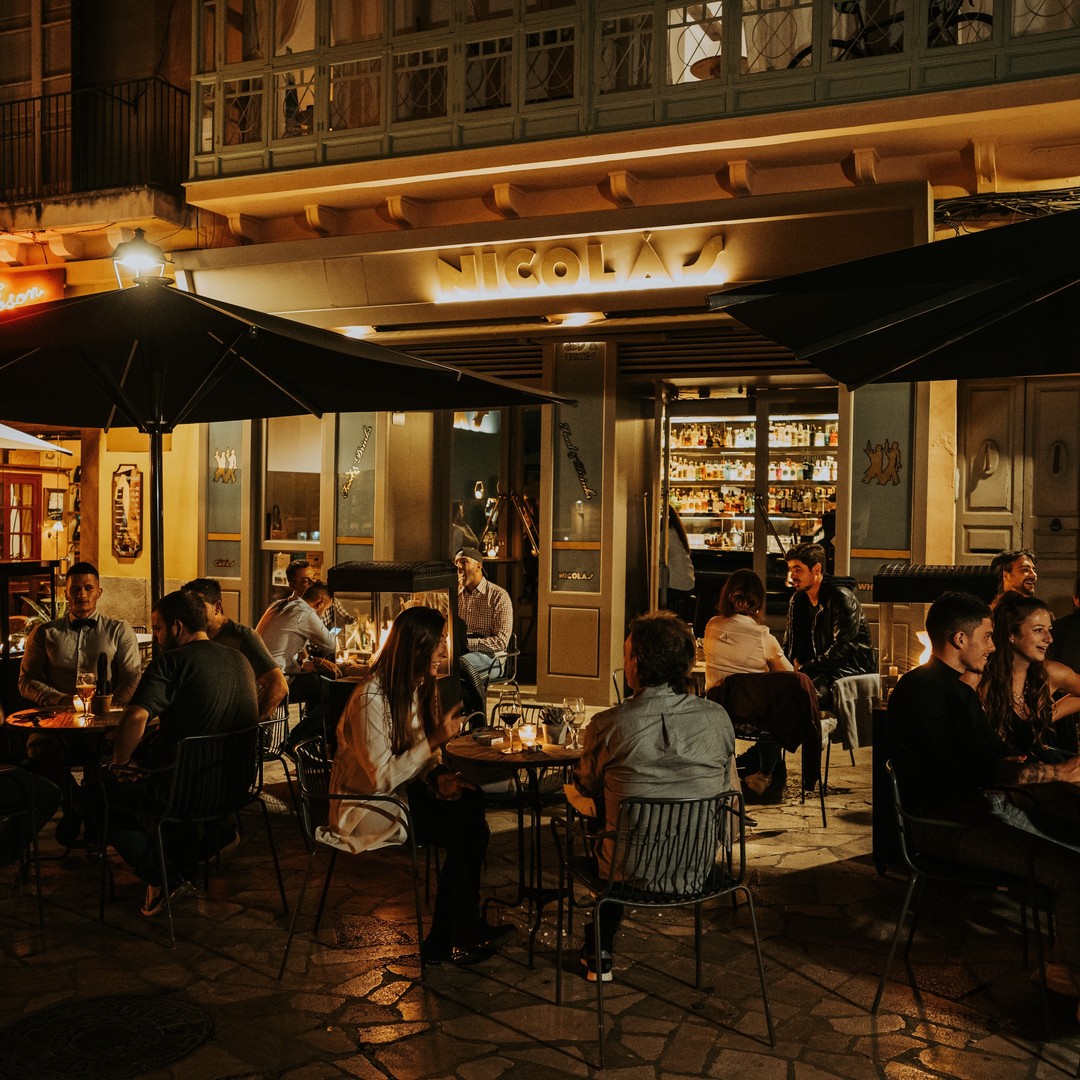 El Bar Nicolás ofrece una velada acogedora al aire libre con mesas iluminadas, donde amigos disfrutan de la noche y un ambiente relajado.