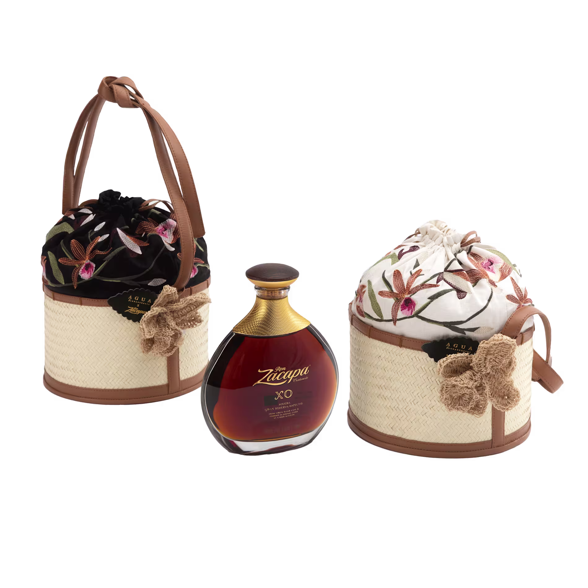 A bottle of Zacapa XO rum is displayed between two woven baskets with floral embroidered fabric lids and brown leather handles on a white background.