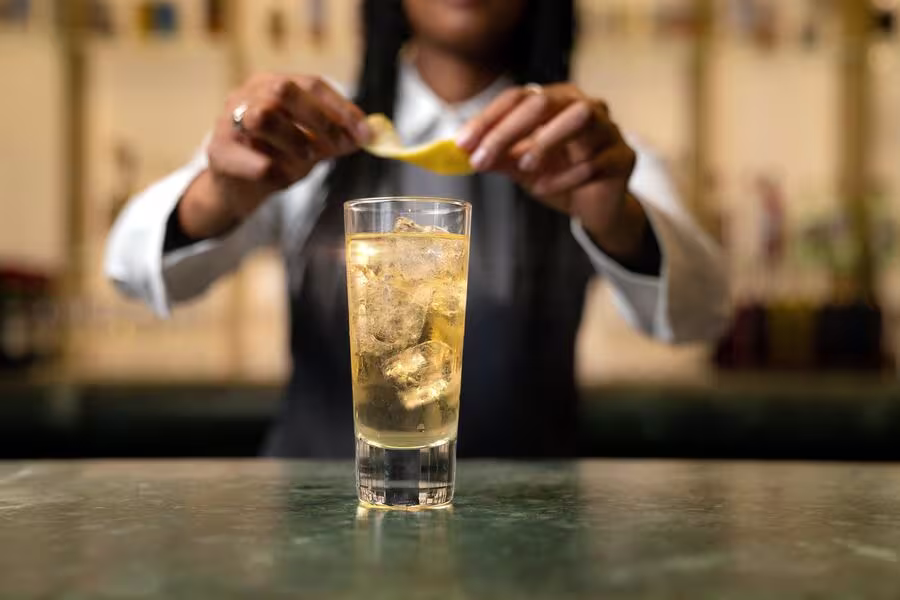 A bartender holds a lemon twist over a tall glass filled with ice and a light-colored drink on a bar counter.