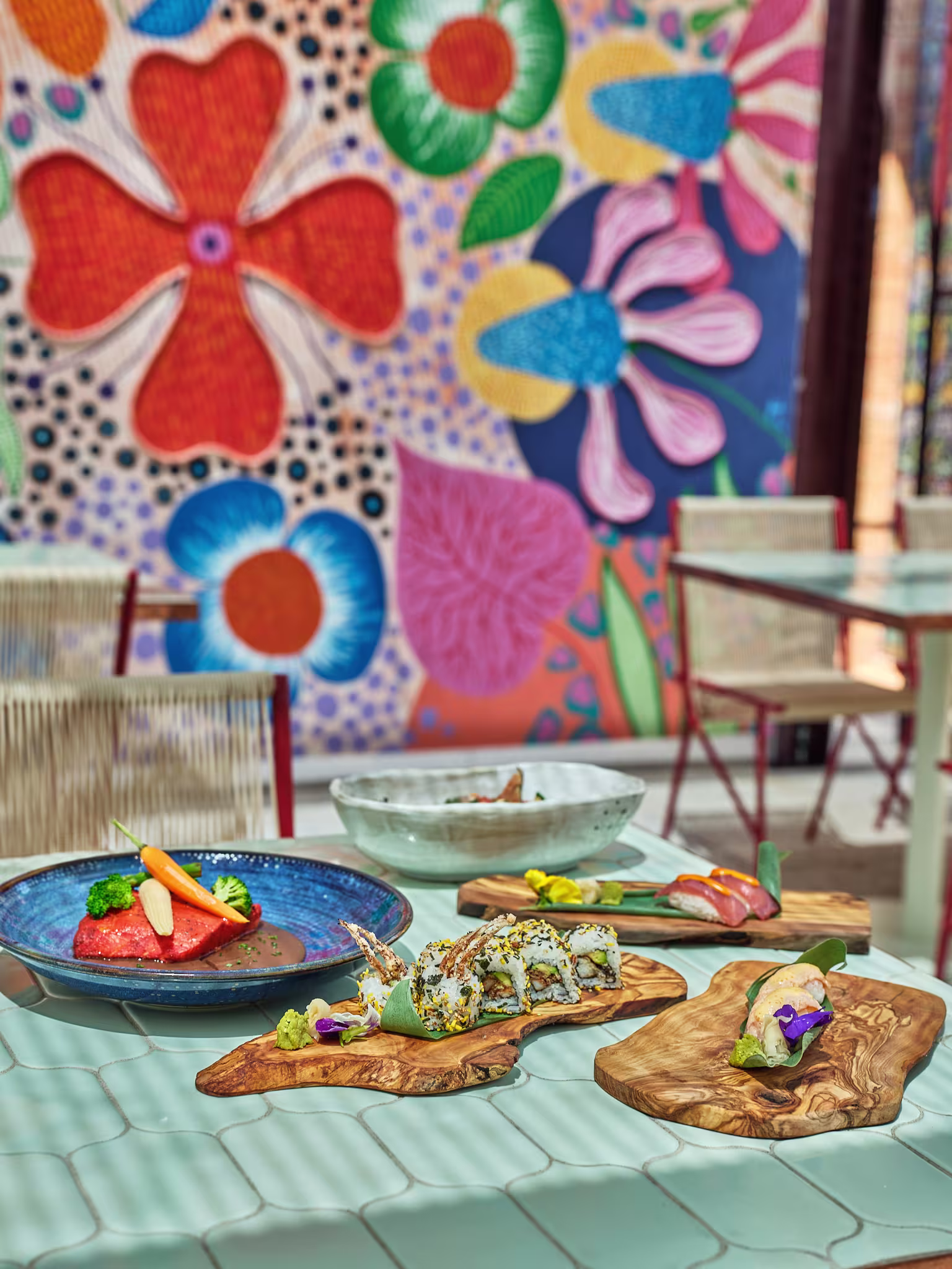 La foto muestra una mesa servida de Torcuato, con varios platos de comida. Hay al menos tres platos con diferentes tipos de alimentos que parecen estar cuidadosamente presentados. En el fondo, hay un mural colorido con patrones florales.