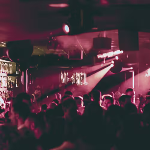 Club lleno de gente bajo luces rosas y rojizas, con un letrero de neón que dice “VESSEL” iluminando el ambiente.