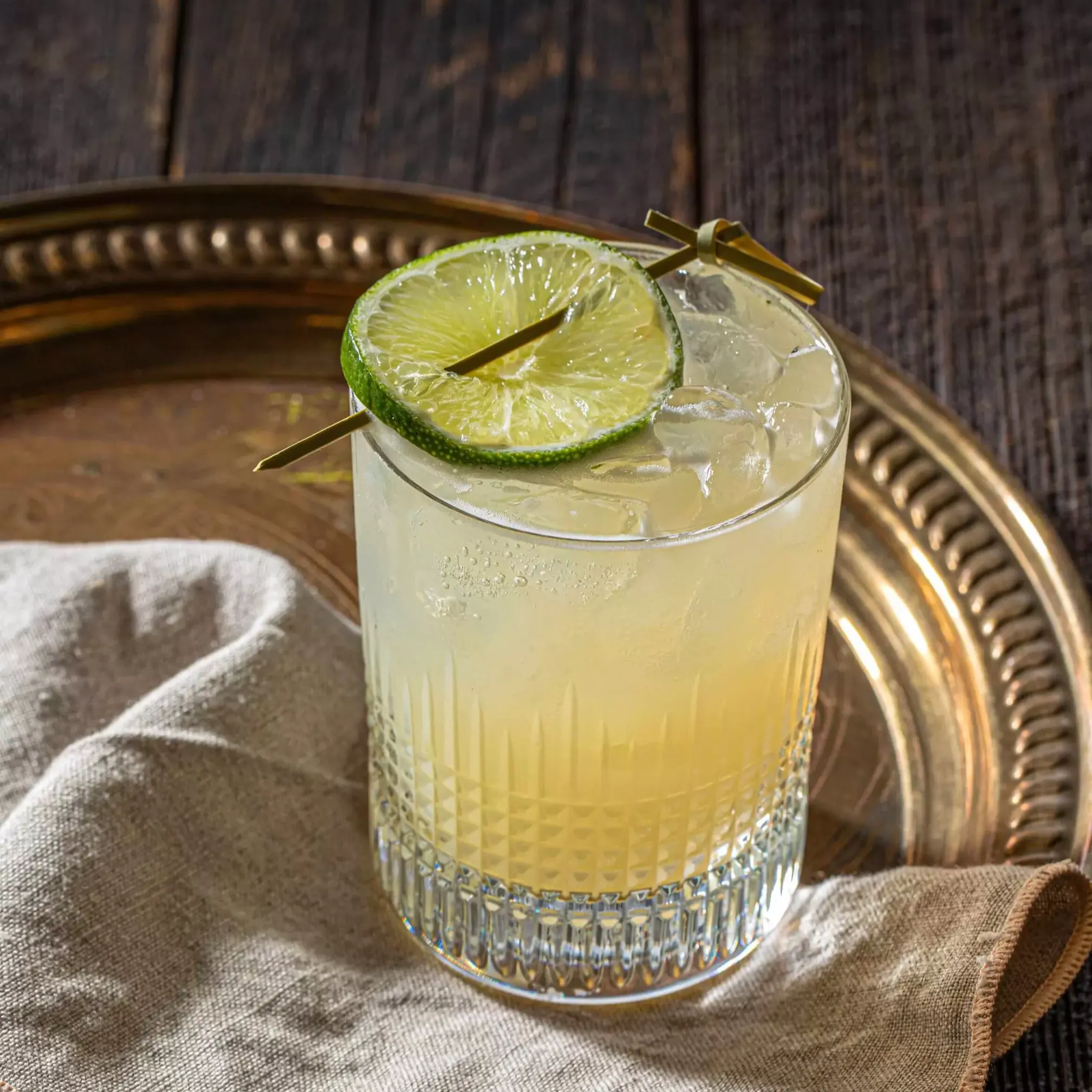 A glass of cocktail with ice and a lime wheel garnish, placed on a cloth napkin atop a round metal tray on a wooden table.