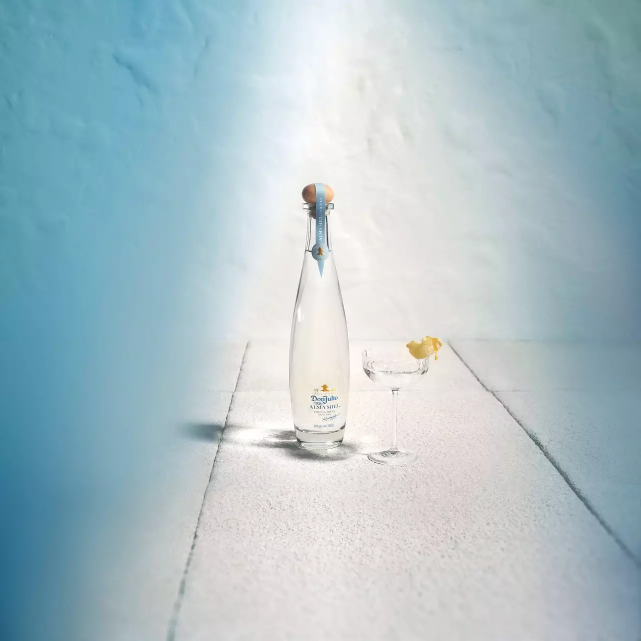 A clear glass bottle of tequila and a coupe glass with a lemon twist sit on a white textured surface against a blue and white background.