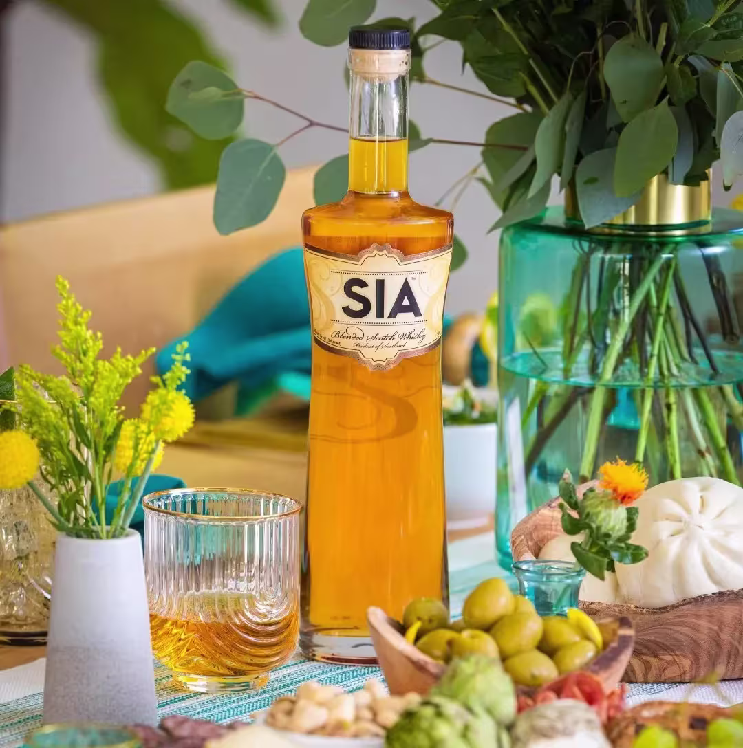 A bottle of SIA blended Scotch whisky stands on a table with a glass of whisky, green olives, bread, and a vase of greenery and yellow flowers.