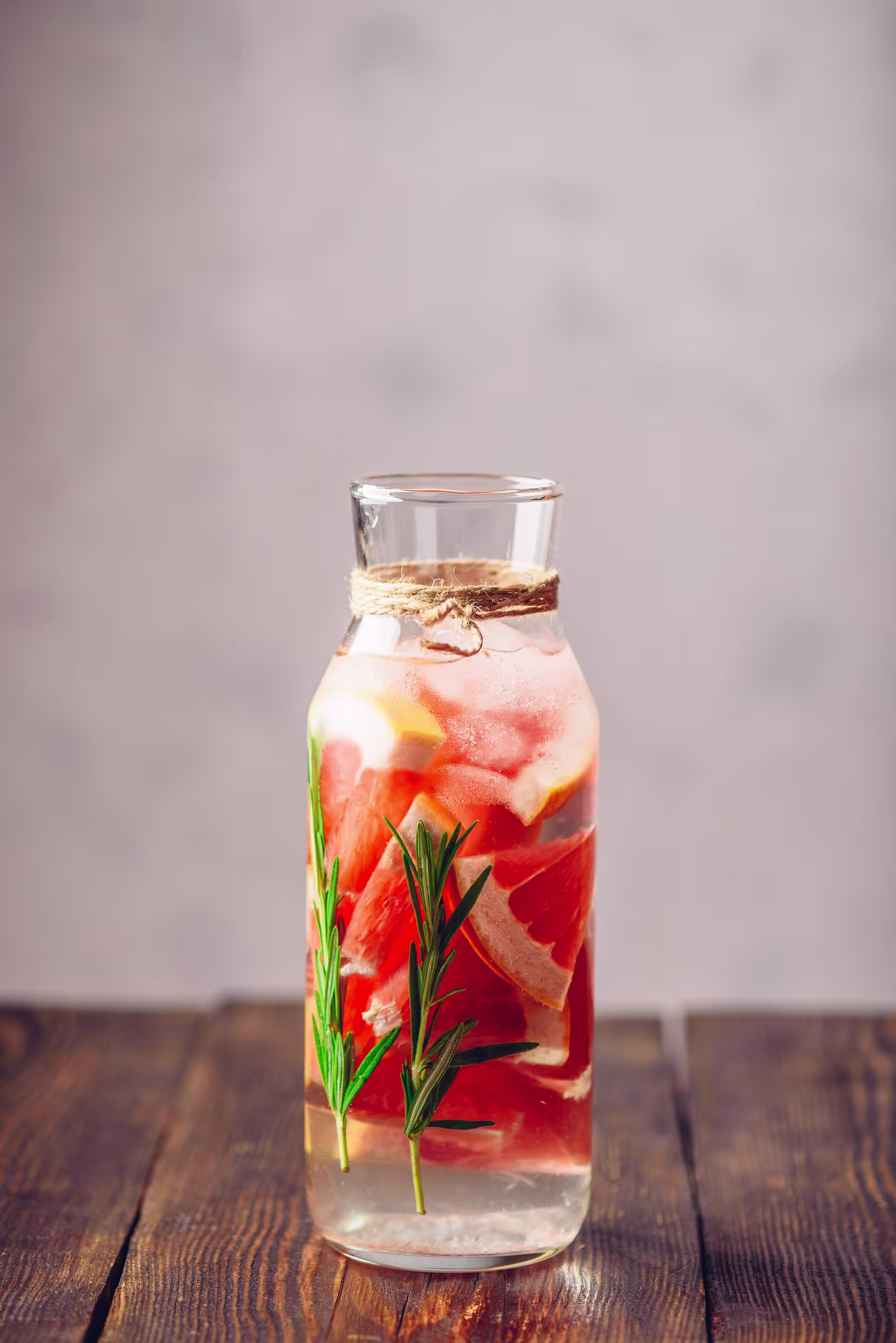 A glass carafe filled with water, grapefruit slices, and sprigs of rosemary, placed on a wooden surface with a neutral background.