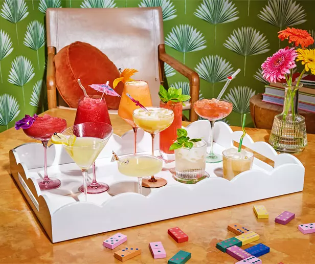 A white tray holds various colorful cocktails in glasses, garnished with fruit and umbrellas, on a table with dominoes; a chair and vase of flowers are in the background.