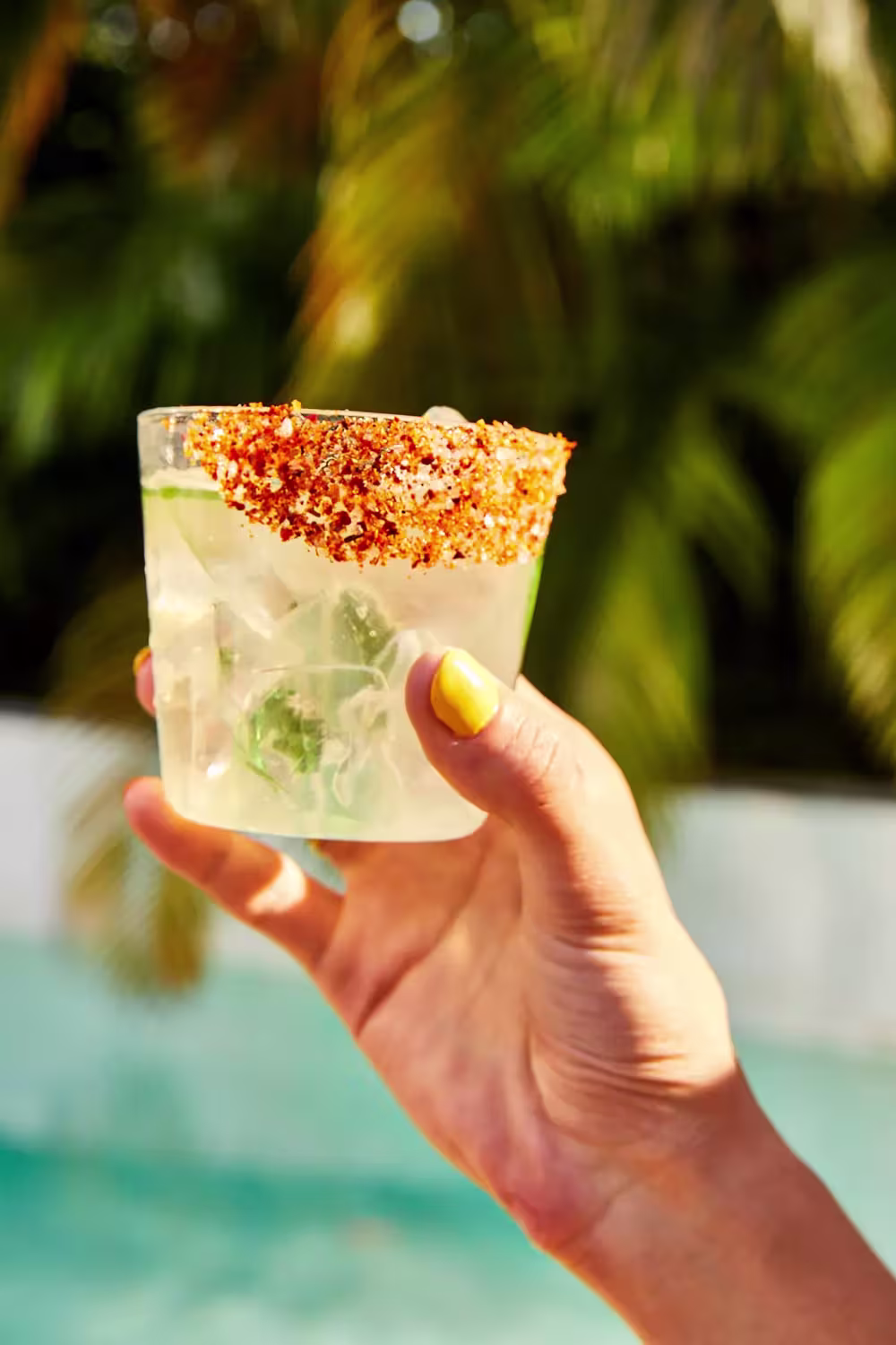 A hand with yellow nail polish holds a glass of iced beverage with a chili powder rim, outdoors near a pool and palm trees.
