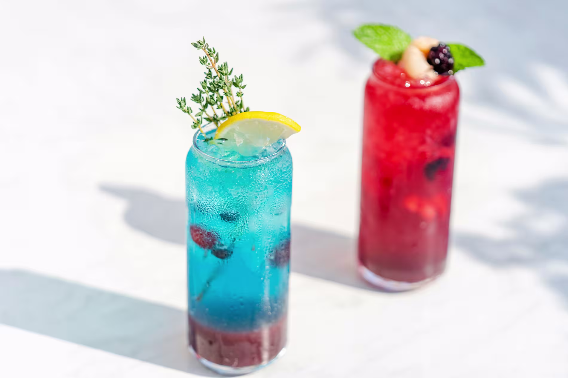Two tall glasses of colorful cocktails on a white surface; one blue with a lemon slice and herbs, the other red with fruit and mint garnish.