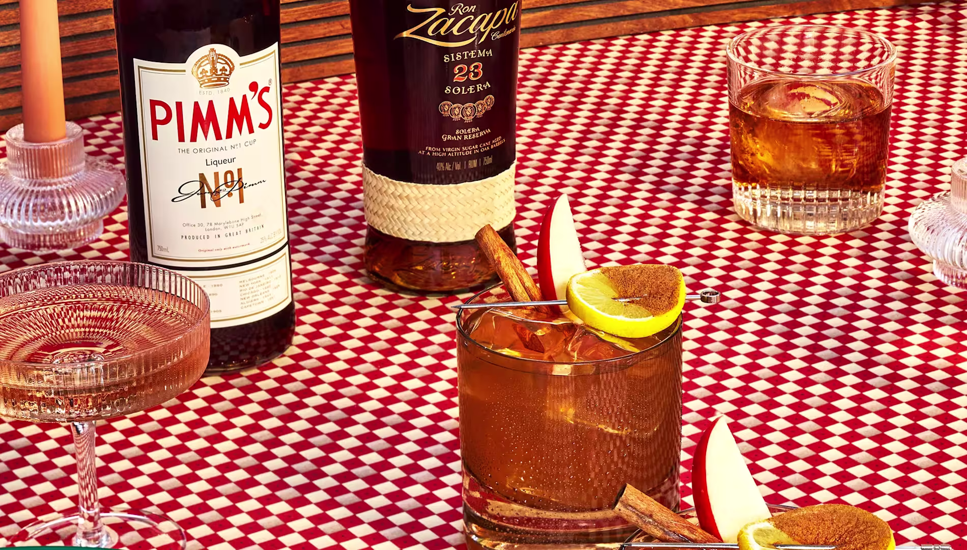 A table with red and white checkered cloth holds bottles of Pimms and rum, cocktails garnished with lemon and cinnamon, a glass of champagne, and candles.