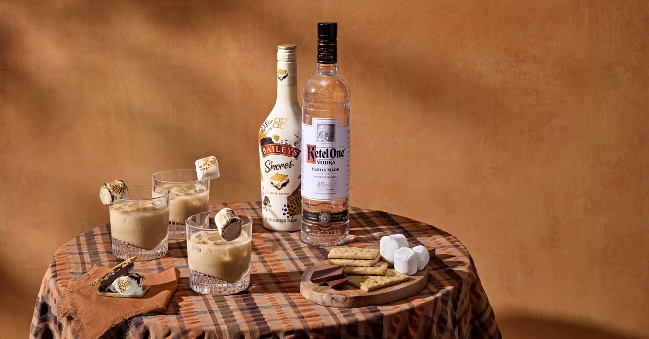 Two bottles of Baileys Smores liqueur and Ketel One vodka sit on a table with three cocktails, toasted marshmallows, crackers, chocolate, and marshmallows against an orange background.