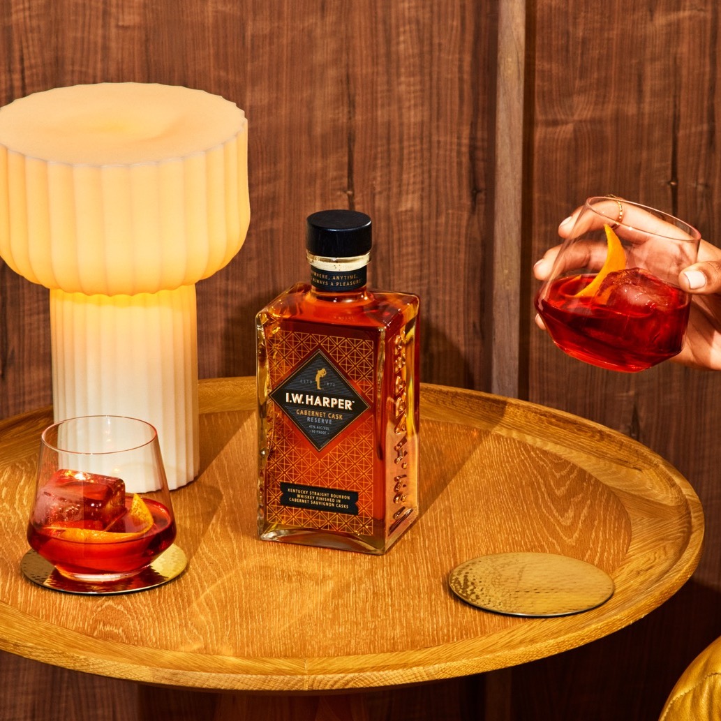 A bottle of I.W. Harper Kentucky bourbon sits on a round wooden table with two glasses of cocktails, a modern lamp, and a gold coaster against a wooden background.