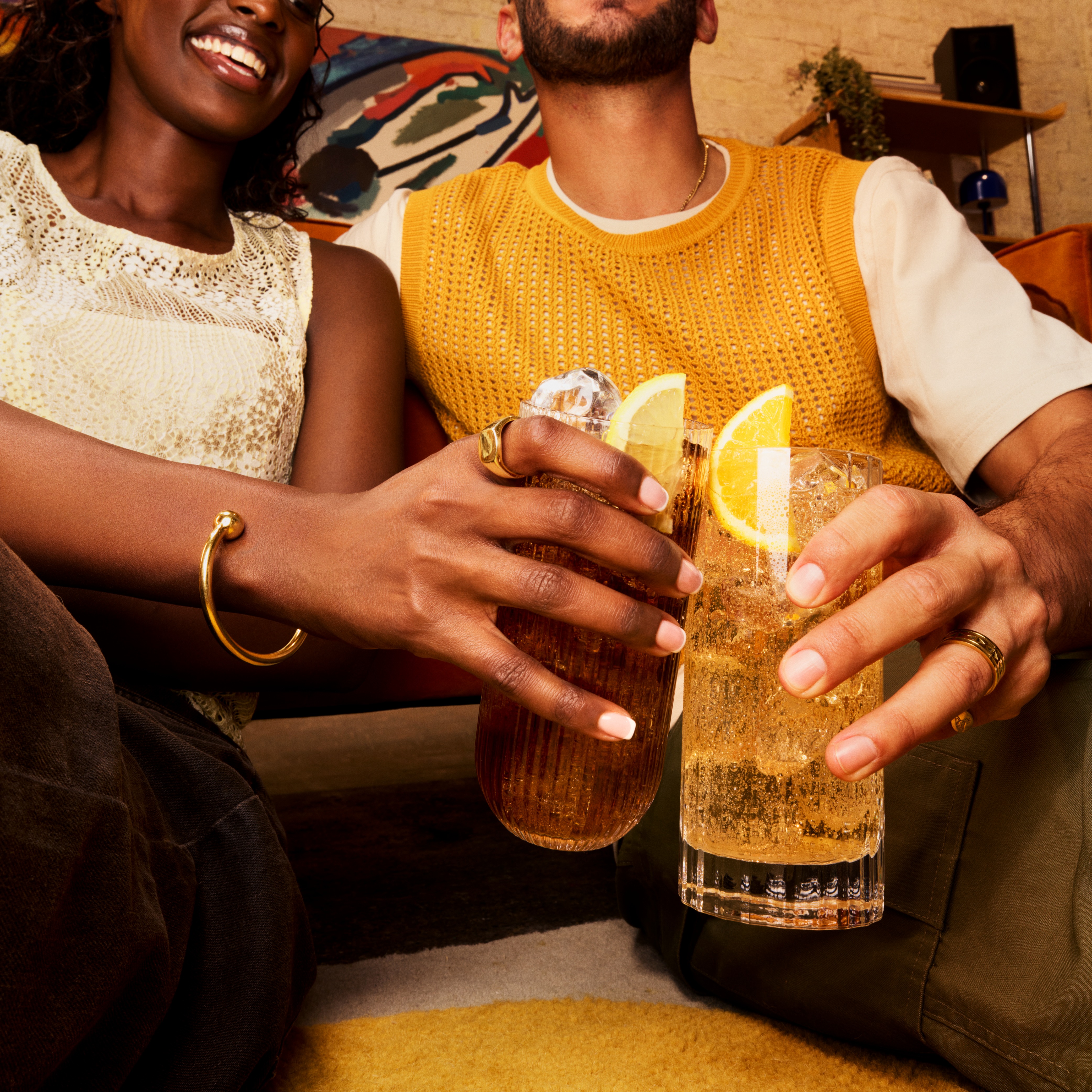 Doi prieteni zâmbitori ciocnesc două cocktailuri „highball” decorate cu felii de lămâie.