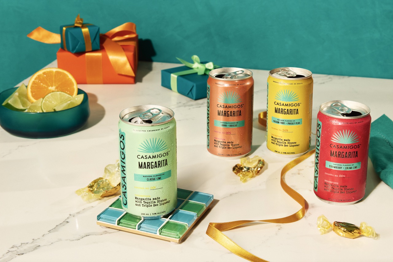 Four cans of Casamigos Margarita are displayed on a marble surface with ribbons, wrapped gifts, candy, and a bowl of sliced citrus fruits.