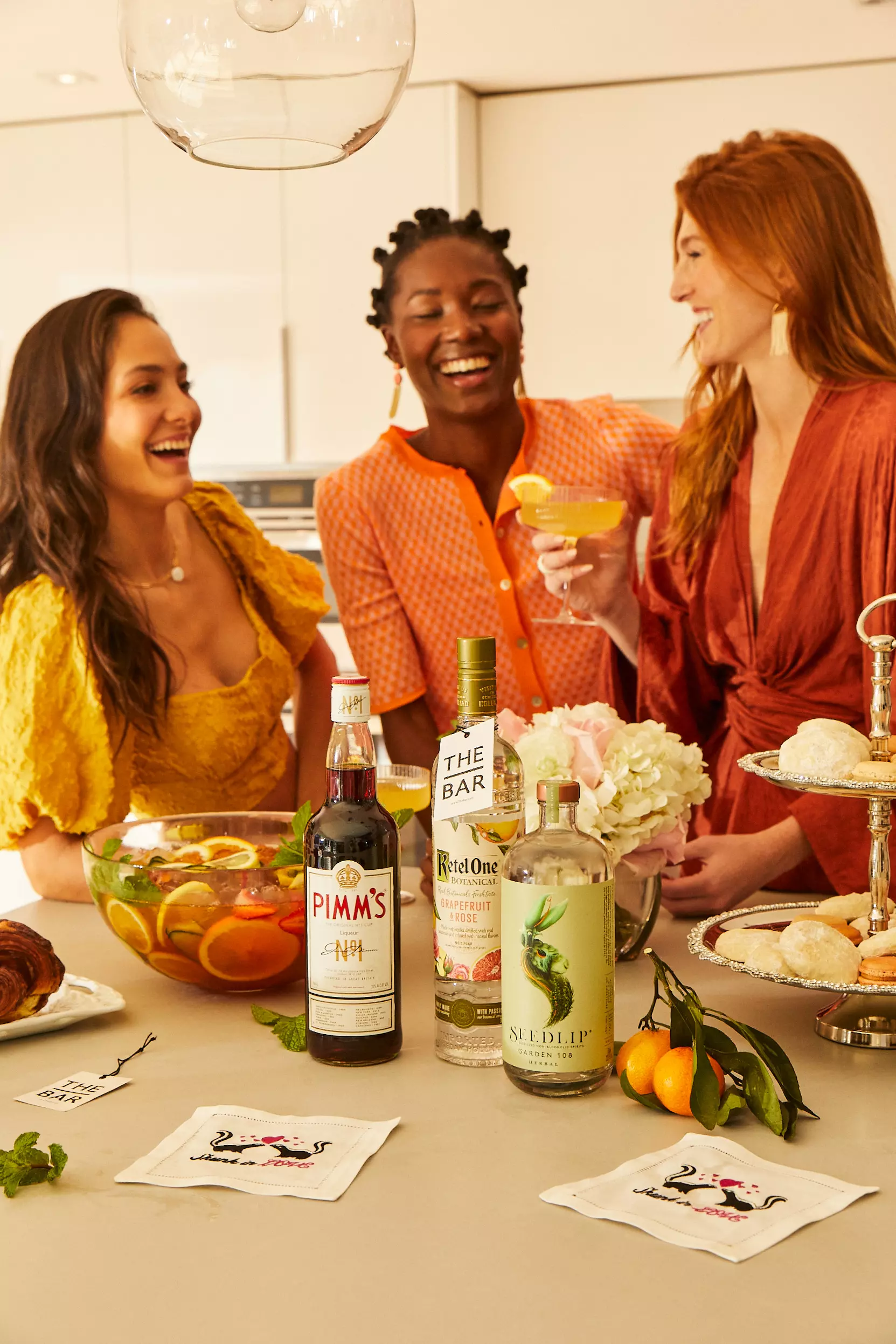 Three women stand around a kitchen counter with cocktails, a punch bowl, bottles of Pimm’s, Ketel One, and Seedlip, snacks, and a floral arrangement, laughing together.