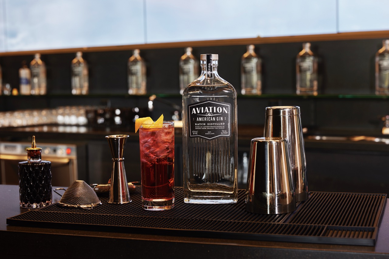 A bottle of Aviation American Gin, a red cocktail with lemon garnish, and bartending tools are arranged on a bar counter.