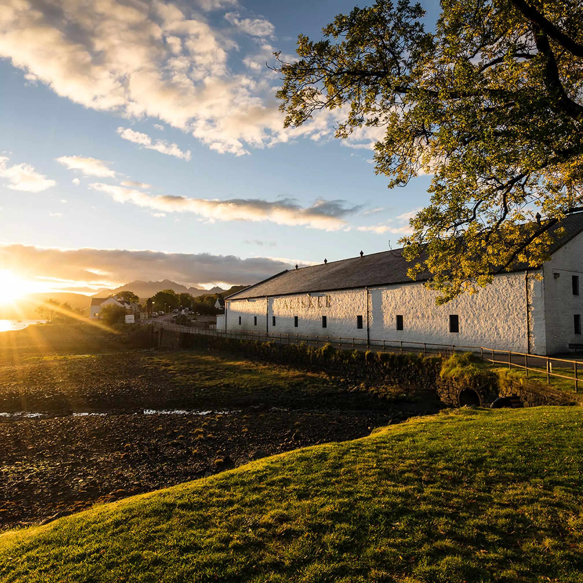 White stone building beside a rocky shoreline at sunset, with sunlight casting long shadows across grass and trees in the foreground.