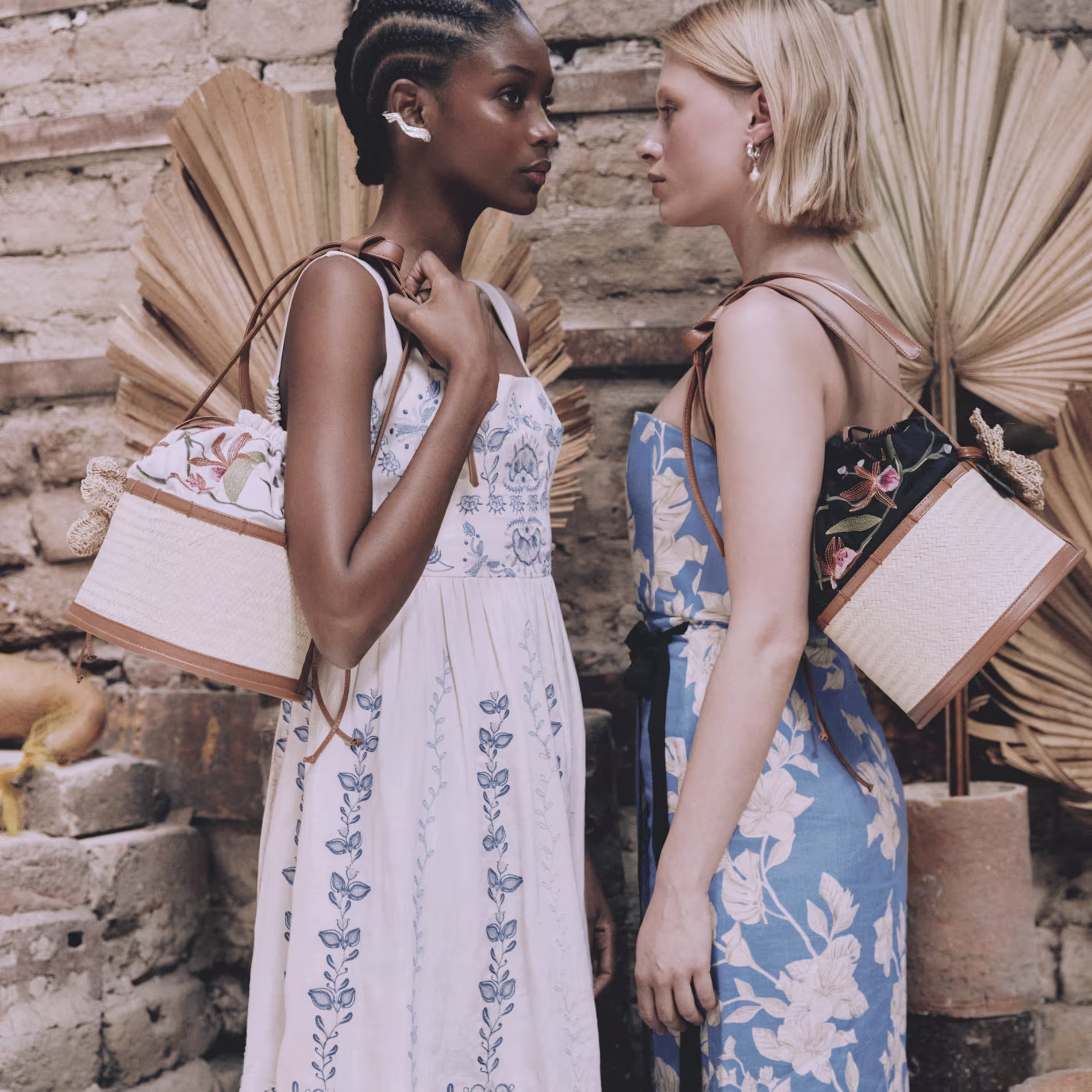 Two women in floral dresses stand face to face, each holding a woven handbag, in front of a rustic stone wall with dried palm leaves in the background.