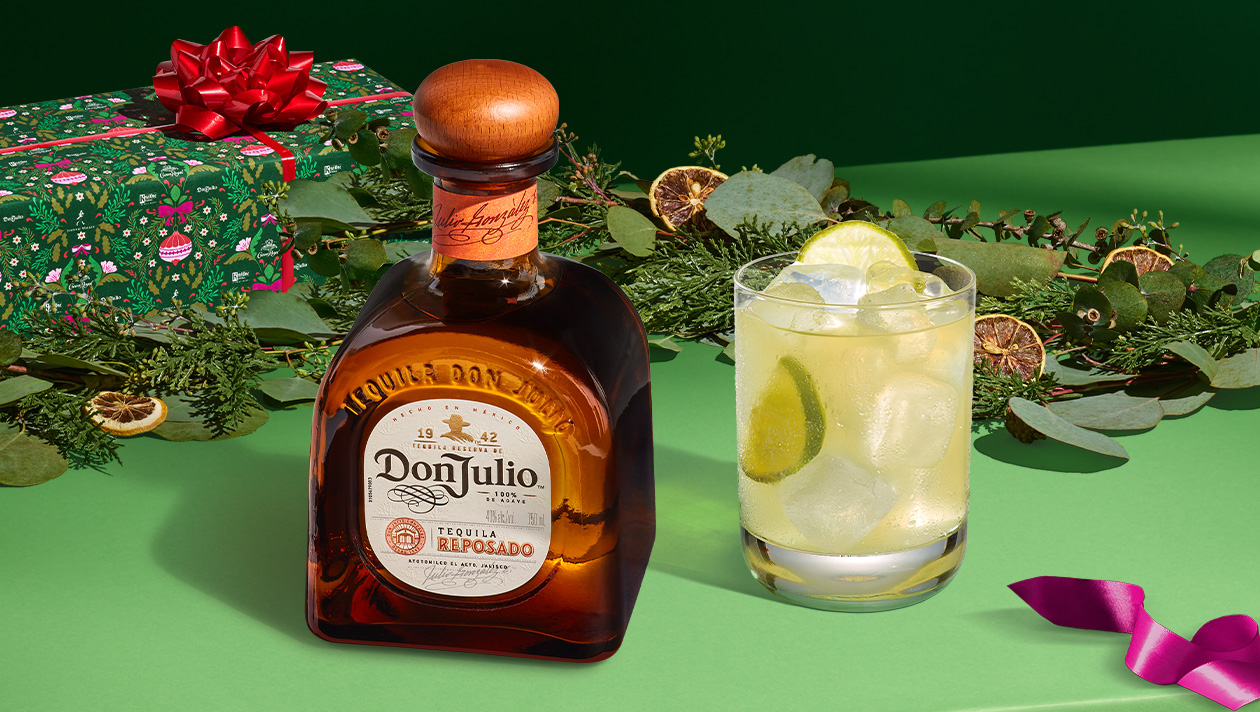 A bottle of Don Julio tequila sits next to a cocktail with ice and lime, surrounded by greenery, a wrapped gift, and holiday decorations on a green surface.