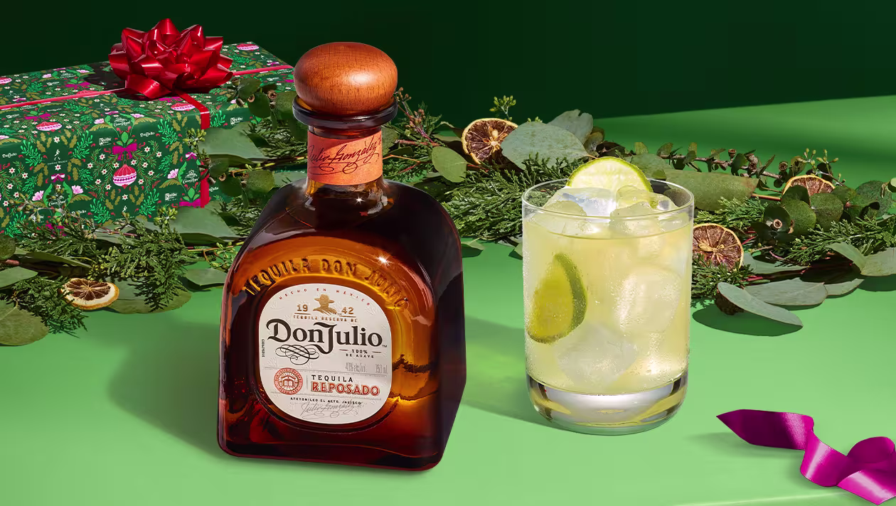 A bottle of Don Julio tequila sits next to a cocktail with ice and lime, surrounded by greenery, a wrapped gift, and holiday decorations on a green surface.