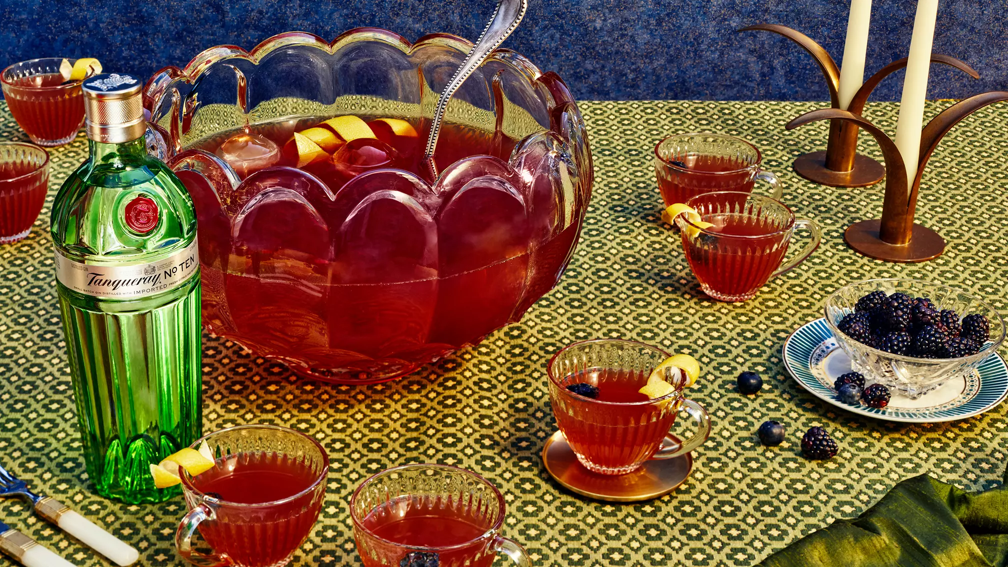 A punch bowl with red punch and lemon slices, surrounded by matching cups, a bottle of Tanqueray gin, blackberries in a dish, and candlesticks on a patterned tablecloth.