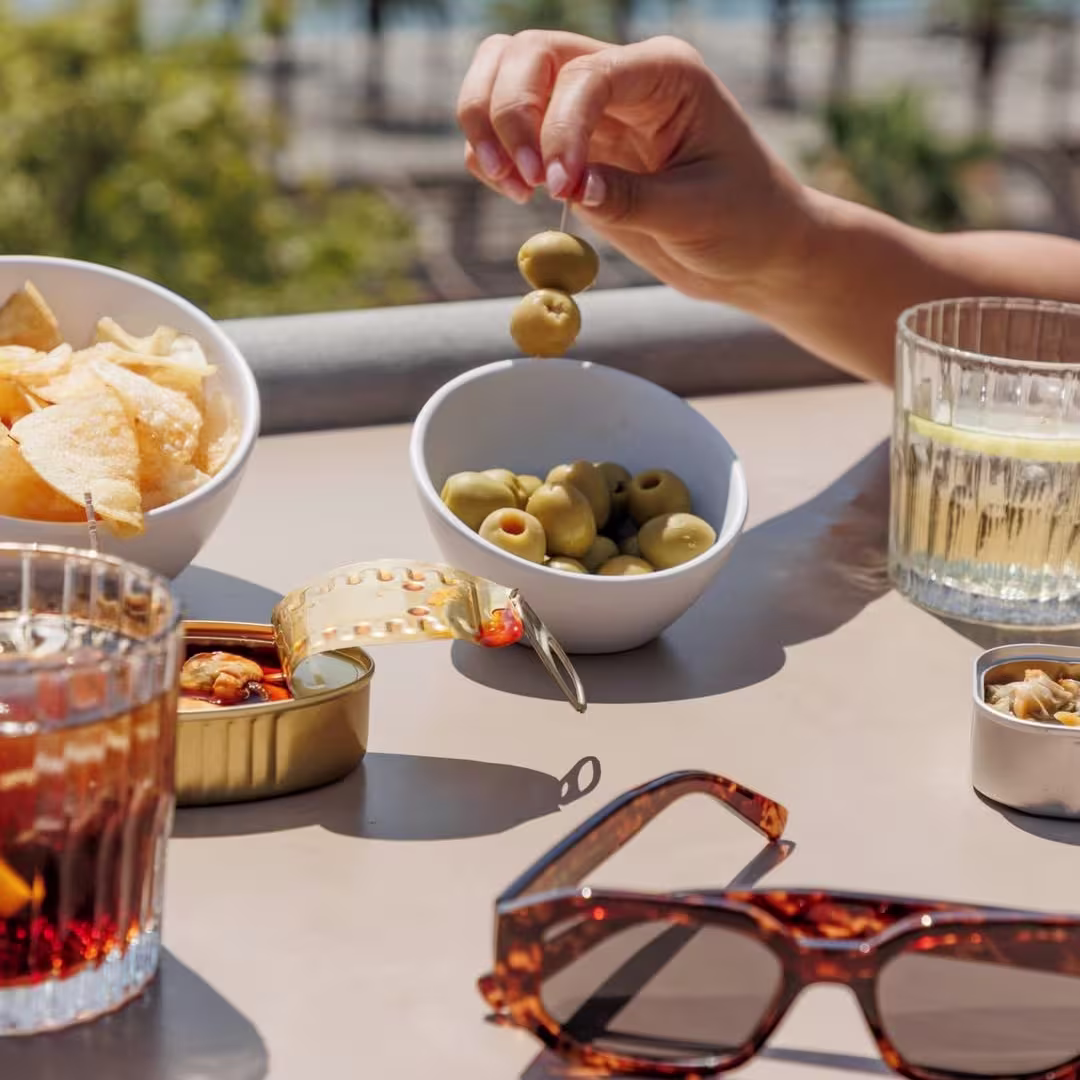 Aperitivo en la terraza de La Duquesa de Cardona con vistas al puerto: vermut, aceitunas, patatas y conservas al sol, junto a unas gafas de sol sobre la mesa.