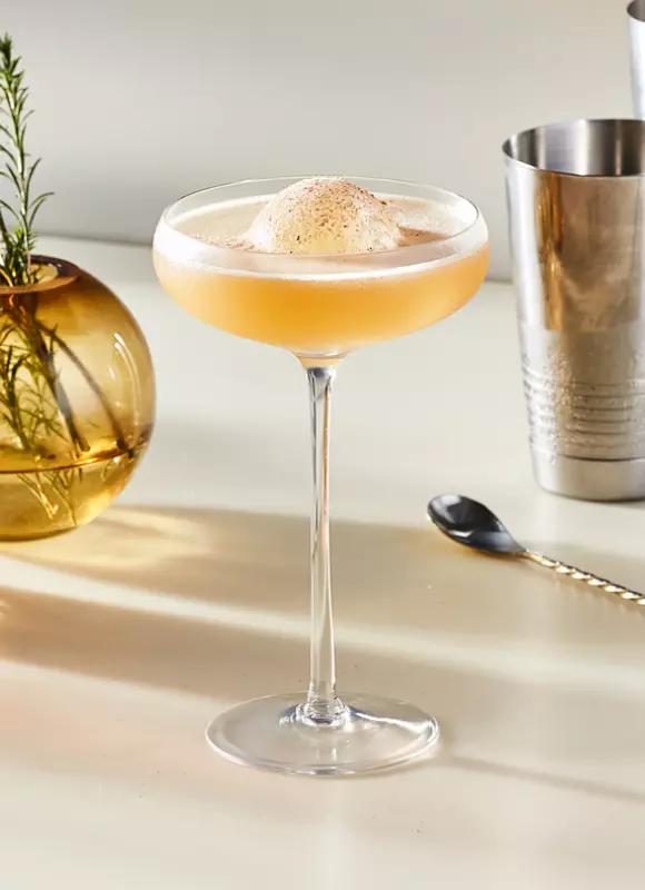 A coupe glass filled with a frothy, light brown cocktail garnished with foam, sits on a light surface next to a cocktail shaker, bar spoon, and a round vase with rosemary.