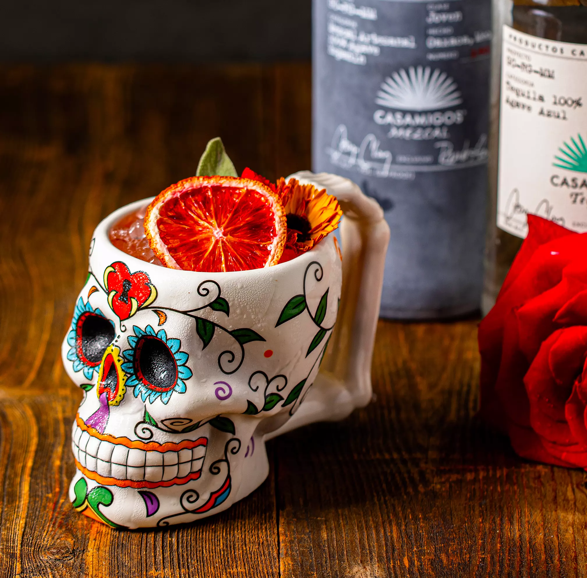A colorful skull-shaped mug filled with a cocktail, garnished with dried orange slices, sits on a wooden surface next to a tequila bottle and a red rose.