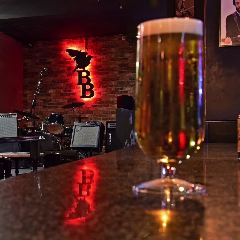 Vista interior de BlackBird, el equipo musical montado en un escenario, lo que indica un lugar para actuaciones de música en vivo. En primer plano, hay una cerveza en la barra del bar. La iluminación es tenue con focos que iluminan el escenario la cerveza