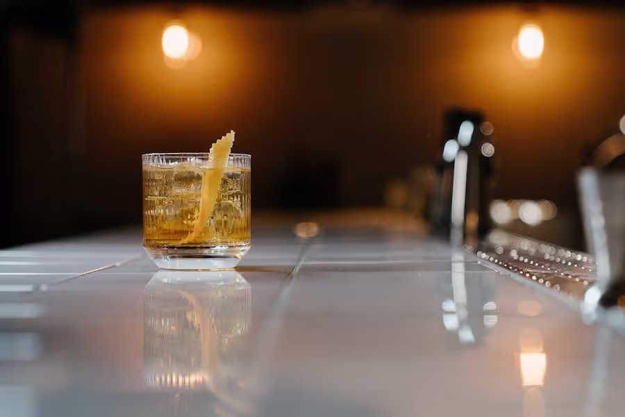 A glass of cocktail with ice and a lemon twist garnish sits on a white tiled bar counter with warm lighting in the background.