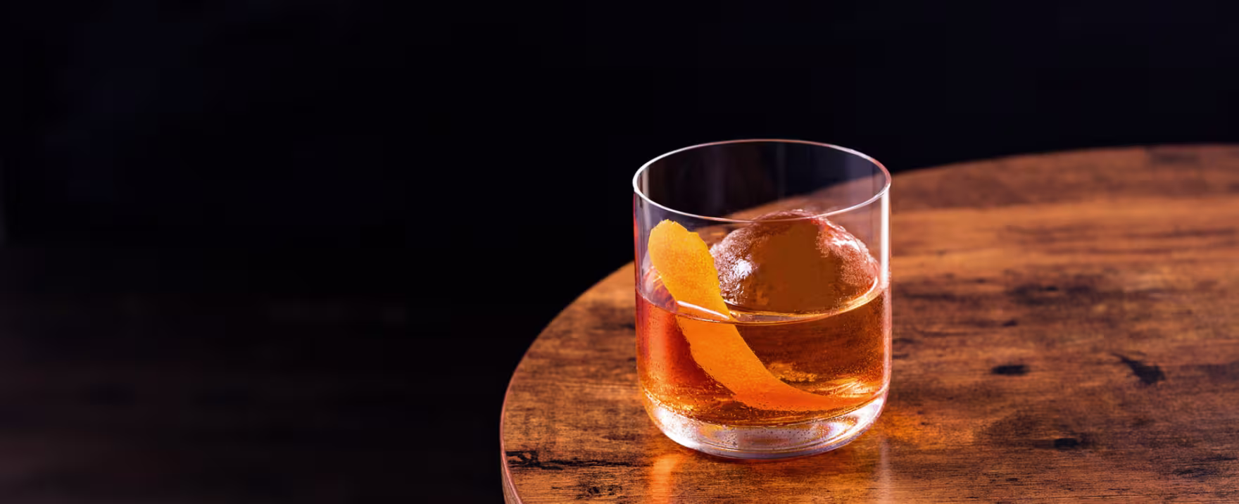 A glass of cocktail with ice and a lemon twist garnish sits on a white tiled bar counter with warm lighting in the background.