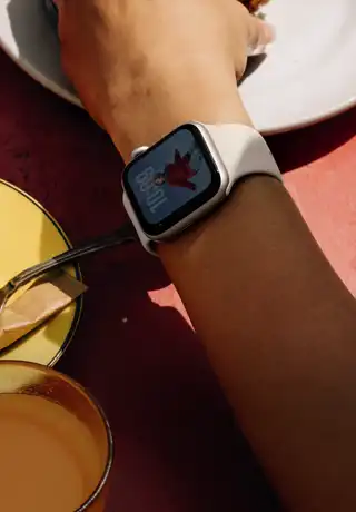 A person's arm grabbing a breakfast food, Apple Watch SE 3 on wrist, Starlight aluminum case, always-on display, portrait of a person on watch face, starlight Sport Band