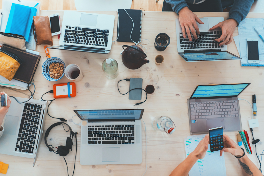Vogelansicht auf einen großen Arbeitstisch auf dem Laptops, kopfhörer Getränke und Hände mit Handys zu sehen sind