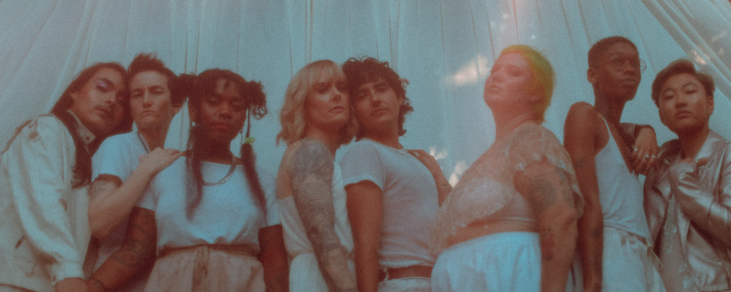 A group of LGBTQIA+ people in white and blush outfits looking directly at the person taking the photo.