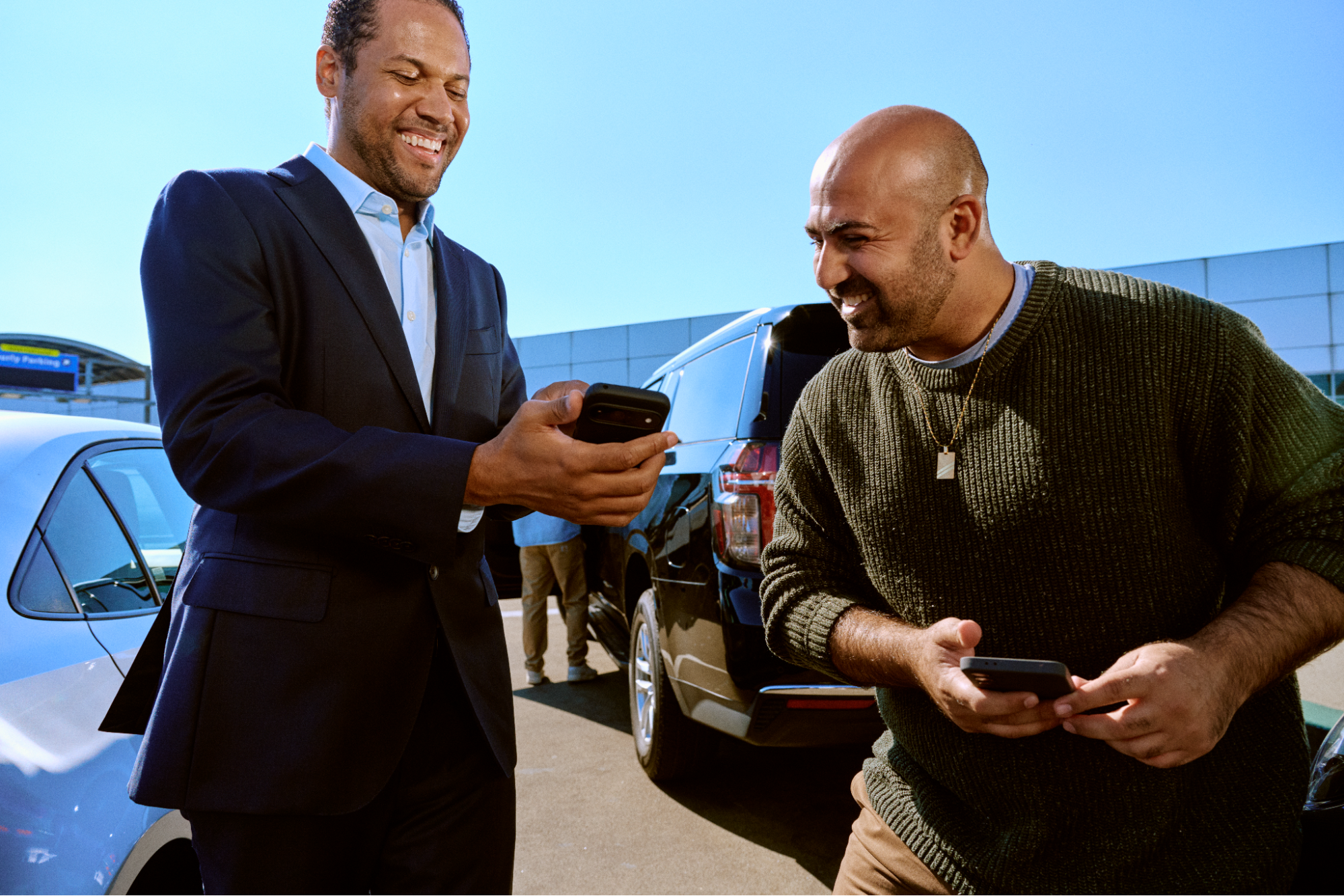 Two smiling men looking at smartphones while standing next to cars under blue sky.
