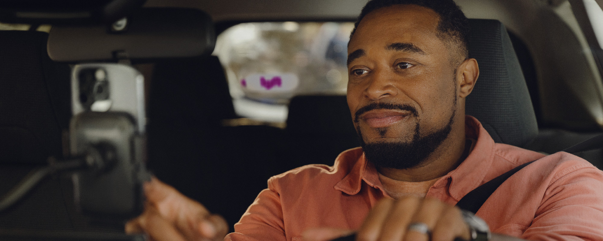 A man in a pink shirt in the driver's seat of a car, reaching toward a phone mounted on the dashboard with a Lyft logo in the background.