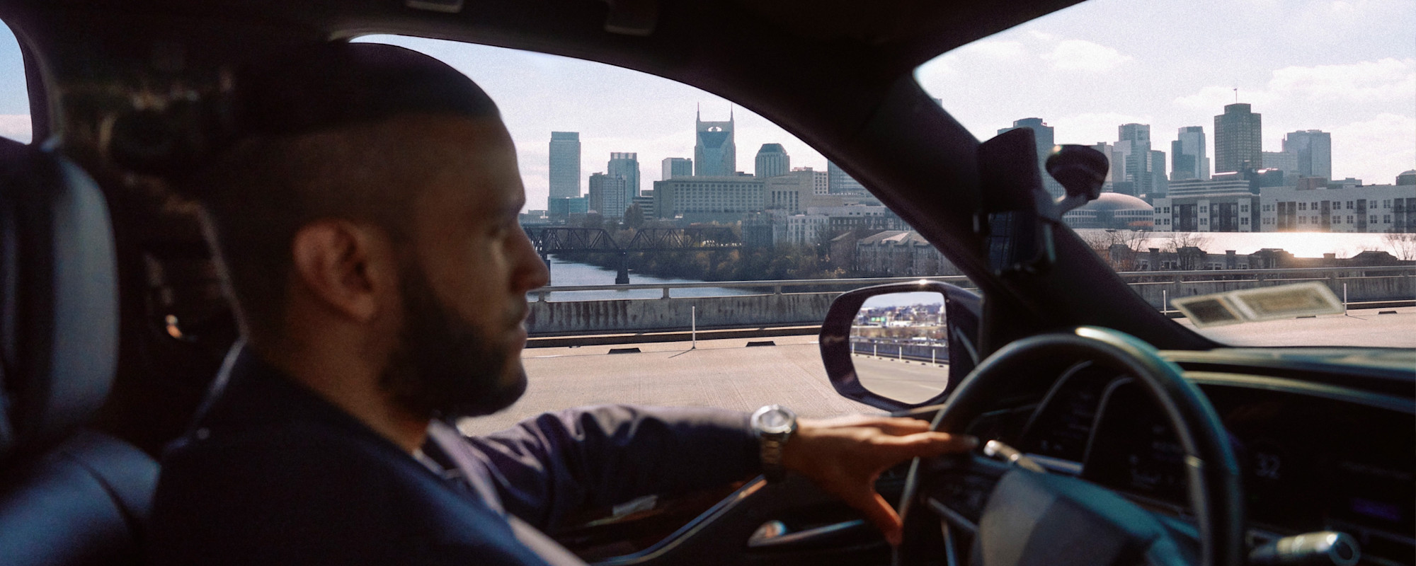 A man with a beard and a kippah drives a car with a city skyline and a river visible through the windshield.